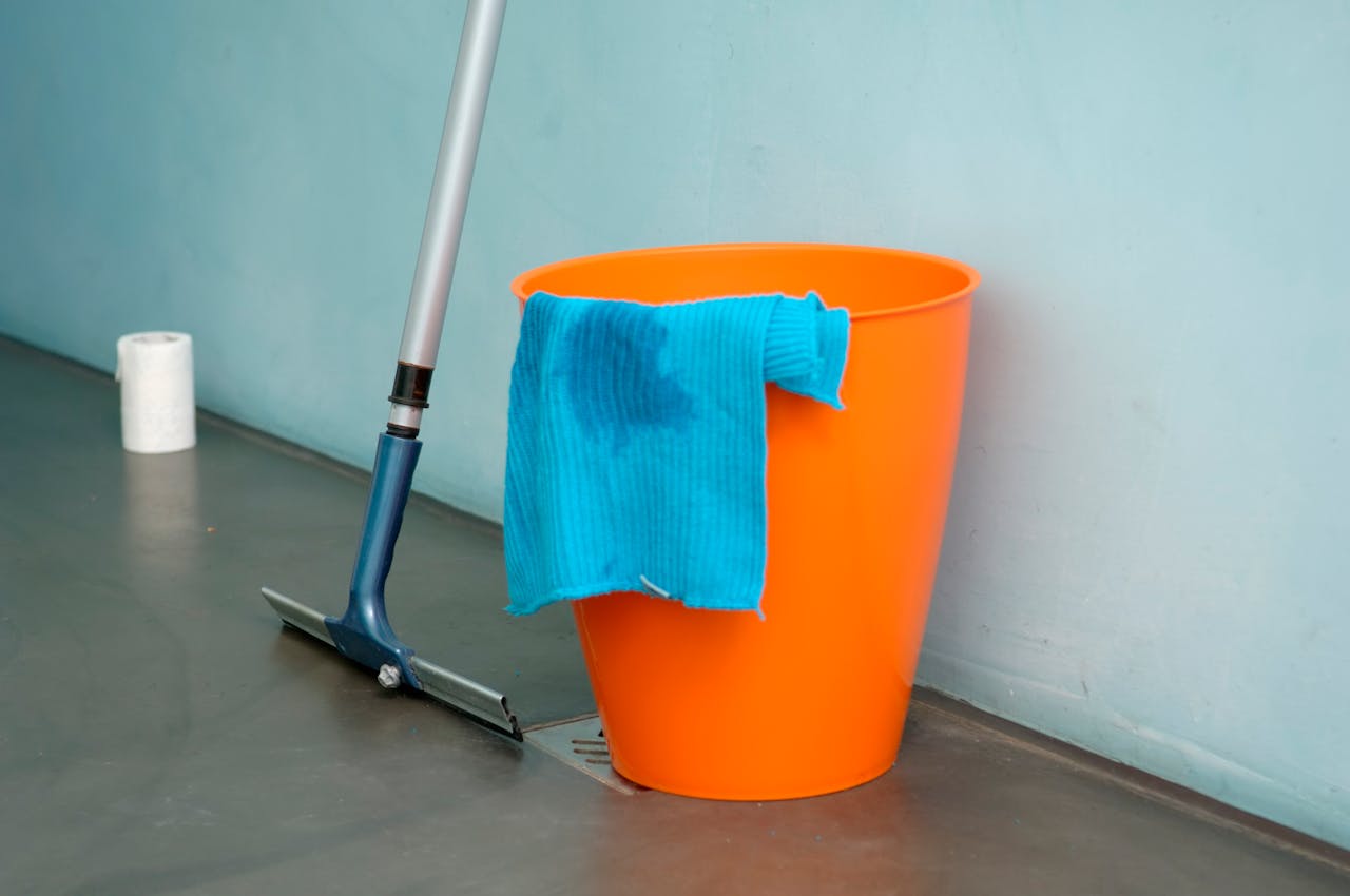 about-02 Close-up of cleaning supplies including orange bucket, mop, and blue rag indoors.